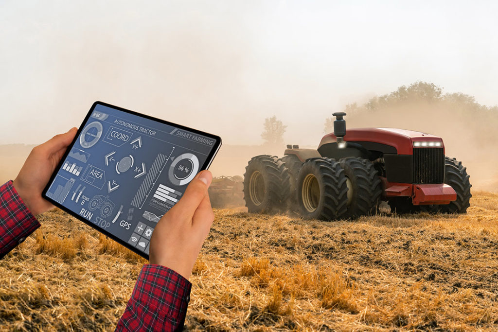 A farmer holding a tablet with a display showing data and controls for smart farming, with a focus on an autonomous tractor operating in the background on a field.