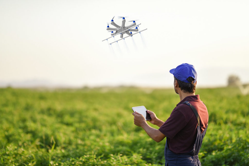 A farmer with a tablet controls an agricultural drone hovering over a green crop field, showcasing modern precision farming technology.
